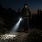 A man hiking on a rocky mountain trail at dusk, holding a flashlight that illuminates the path. He is wearing an olive green jacket and dark pants, with pine trees and mountains in the background under a twilight sky.